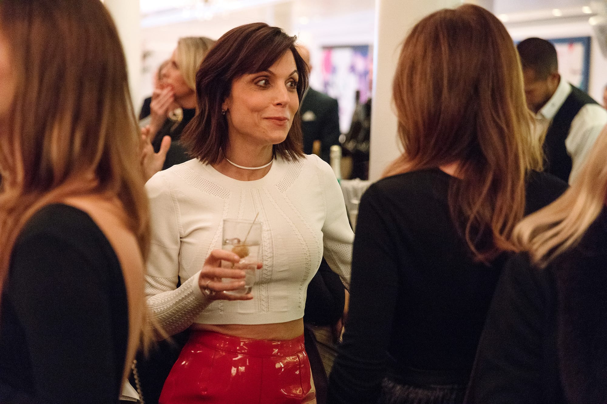 Bethenny Frankel, wearing a white top and red pants, holding a drink and talking to another woman at an event.