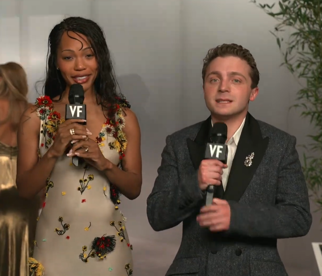 Two people hold microphones on a red carpet; one wears a floral dress, the other a dark suit with a decorative brooch