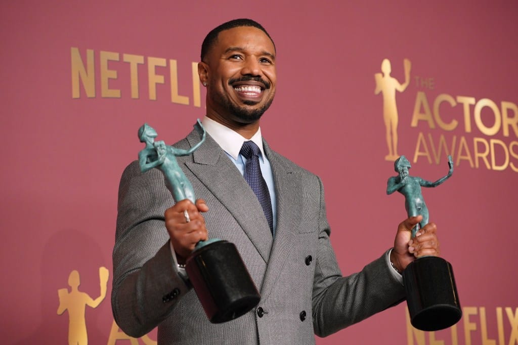 Michael B. Jordan, in a grey suit and blue tie, smiles holding two SAG Awards, recognized for outstanding performance in "Sinners."