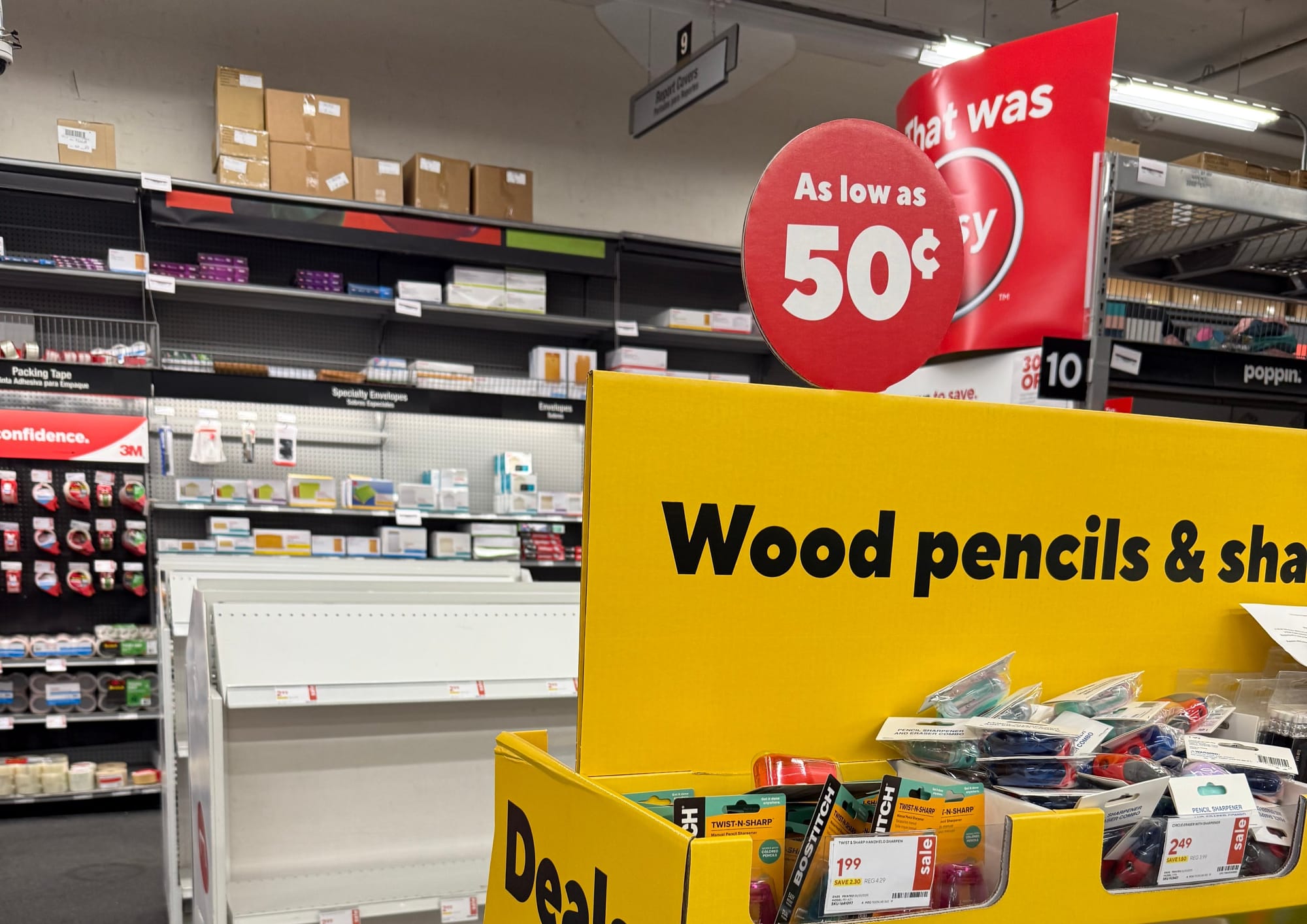 Store aisle with school supplies, featuring a prominent sale sign for 50% off. Yellow display stand holds pencils and sharpeners on sale